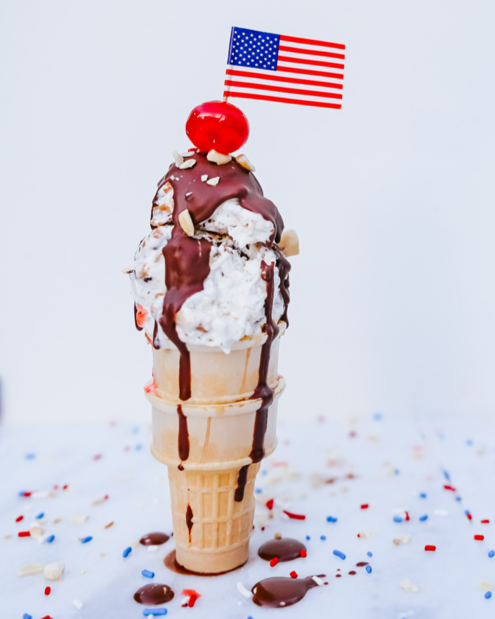 Teddie Peanut Butter ice cream in a cone with chocolate sauce that is topped with a cherry and an American Flag toothpick.  